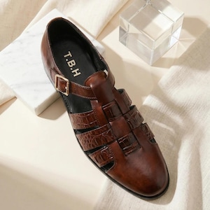 May include: A dark brown leather fisherman sandal with a crocodile-embossed texture, a closed rounded toe, and a gold-tone metal buckle strap. The interior footbed is black and features the white text T.B.H printed near the heel area.
