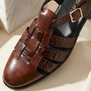 May include: A dark brown leather fisherman sandal featuring a glossy crocodile-embossed texture on the woven straps, a rounded toe, and a gold-toned metal buckle closure on the side strap. The interior footbed is black with partial text visible.