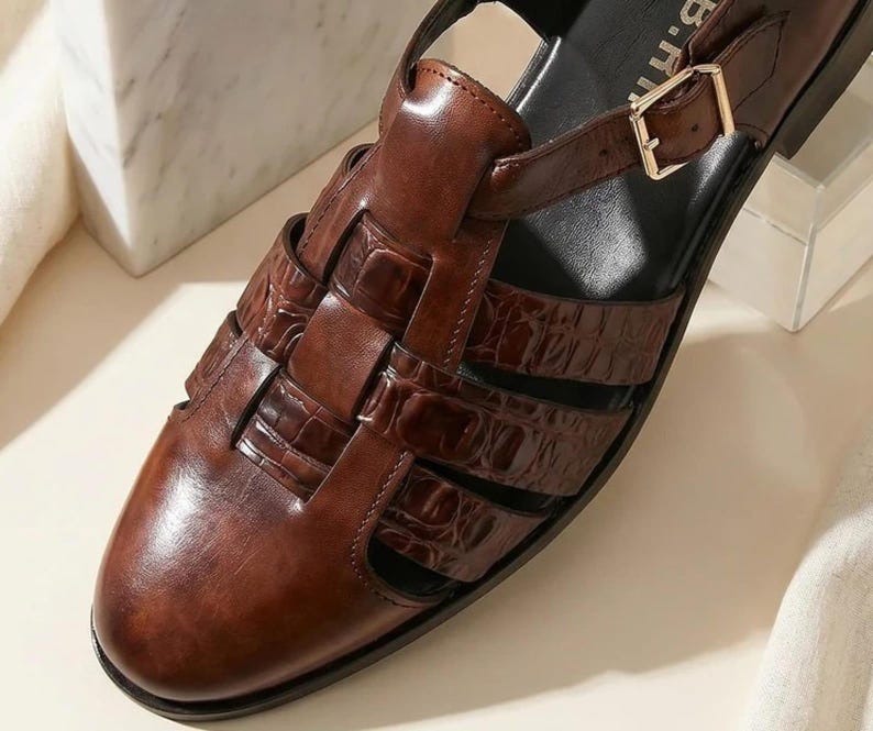 May include: A dark brown leather fisherman sandal featuring a glossy crocodile-embossed texture on the woven straps, a rounded toe, and a gold-toned metal buckle closure on the side strap. The interior footbed is black with partial text visible.