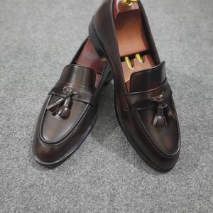 May include: A pair of dark brown leather tassel loafers featuring a smooth finish, a rounded toe, and a decorative strap across the vamp with two tassels. Each shoe contains a wooden shoe tree with a gold-colored knob at the heel.