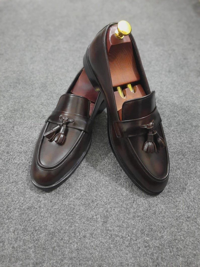 May include: A pair of dark brown leather tassel loafers featuring a smooth finish, a rounded toe, and a decorative strap across the vamp with two tassels. Each shoe contains a wooden shoe tree with a gold-colored knob at the heel.