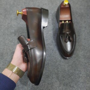 May include: A pair of dark brown leather loafers featuring a tassel detail on the vamp, a rounded toe, and a stacked heel. Each shoe includes a wooden shoe tree with a gold-toned metal knob inserted inside to maintain the shape of the footwear.