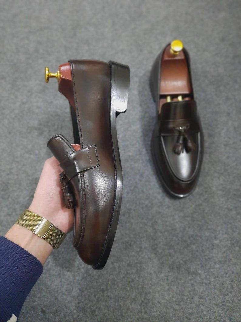 May include: A pair of dark brown leather loafers featuring a tassel detail on the vamp, a rounded toe, and a stacked heel. Each shoe includes a wooden shoe tree with a gold-toned metal knob inserted inside to maintain the shape of the footwear.