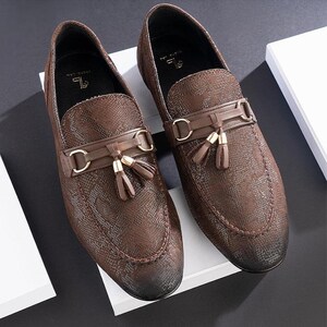 May include: A pair of brown leather loafers with a textured pattern. The shoes feature a decorative strap with gold-colored hardware and small tassels. The shoes are displayed on a white surface.