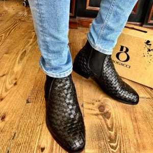 Op de afbeelding: Zwarte leren enkellaarsjes met een geweven design. De laarzen hebben elastische zijpanelen en een puntige neus. De laarzen worden gedragen met een blauwe jeans. Een kartonnen doos met het woord "BOGACCI" staat op de achtergrond.