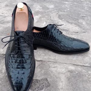 K&ouml;nnte beinhalten: Ein Paar schwarze Lederschuhe mit Krokodilhautstruktur. Die Schuhe haben Schn&uuml;rsenkel und eine spitze Zehenpartie. Die Innenseite des Schuhs ist hellbraun. Die Schuhe stehen auf einer grauen Oberfl&auml;che.