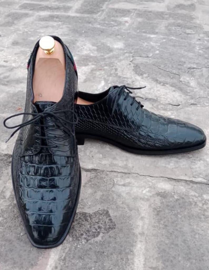 K&ouml;nnte beinhalten: Ein Paar schwarze Lederschuhe mit Krokodilhautstruktur. Die Schuhe haben Schn&uuml;rsenkel und eine spitze Zehenpartie. Die Innenseite des Schuhs ist hellbraun. Die Schuhe stehen auf einer grauen Oberfl&auml;che.