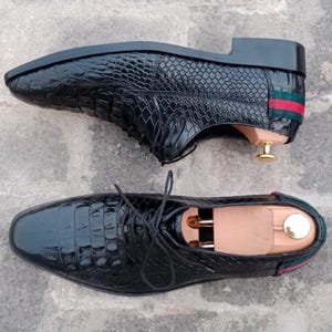 K&ouml;nnte beinhalten: Zwei schwarze Lederschuhe mit Krokodilleder-Textur. Die Schuhe haben ein klassisches Schn&uuml;rdesign und einen kleinen Absatz. Die R&uuml;ckseite der Schuhe hat ein gr&uuml;n-rotes Streifendetail.