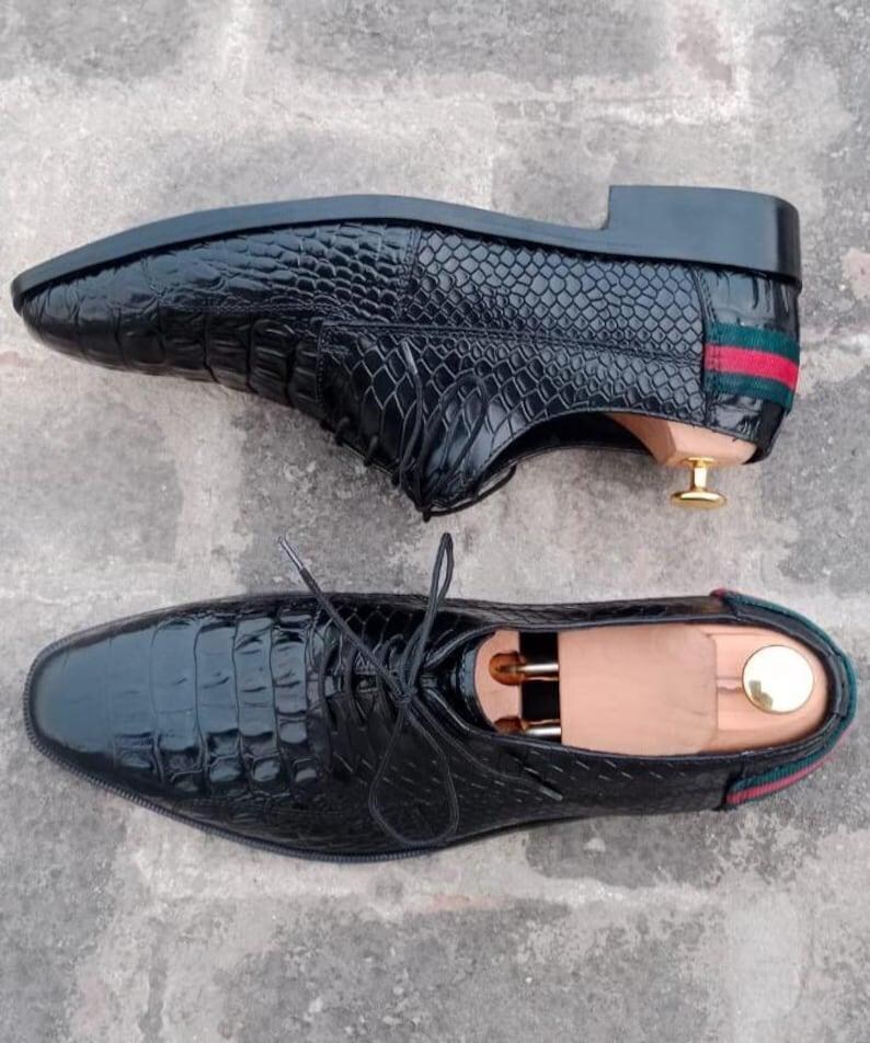K&ouml;nnte beinhalten: Zwei schwarze Lederschuhe mit Krokodilleder-Textur. Die Schuhe haben ein klassisches Schn&uuml;rdesign und einen kleinen Absatz. Die R&uuml;ckseite der Schuhe hat ein gr&uuml;n-rotes Streifendetail.