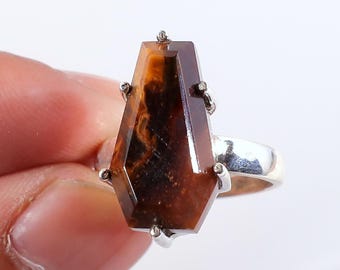 Op de afbeelding: Zilveren ring met een grote, doodskistvormige edelsteen in bruine en amberkleurige tinten. De edelsteen is gezet met kleine zilveren pinnen. De ring staat tegen een witte achtergrond.