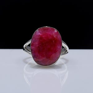 May include: A silver ring with a large, oval-shaped ruby gemstone. The ruby is a rich, deep red hue and dominates the design. The ring's band is a simple, polished silver, showcasing the gemstone.