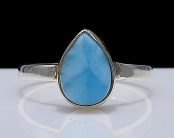May include: A silver ring with a light blue, pear-shaped gemstone. The gemstone is set in a silver band, and the ring is shown on a reflective surface. The ring's design is simple and elegant, suitable for everyday wear.