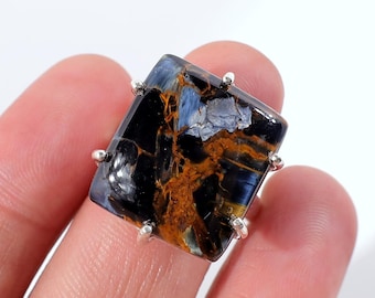 Op de afbeelding: Een vierkante edelsteenring met een gepolijste, veelkleurige steen. De steen vertoont een mix van donkerblauwe, zwarte en goudbruine tinten. De ringzetting is zilverkleurig, met kleine pootjes die de steen vastzetten.
