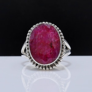 May include: A silver ring featuring a large, oval-cut ruby gemstone. The ruby is a deep, vibrant red color and is set in a bezel setting. The ring has a detailed, beaded border around the gemstone and a split shank design.