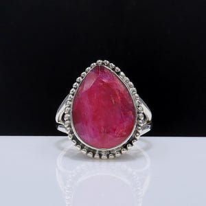 May include: A sterling silver ring featuring a large, pear-shaped, faceted ruby gemstone. The ruby is a vibrant shade of red, set in a bezel with a beaded border. The ring's band has a split design.