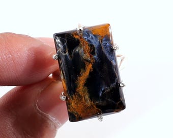 May include: A rectangular gemstone ring featuring a polished, multi-colored stone. The stone displays a mix of dark blue, orange, and brown hues. The ring's setting is silver-toned, with small decorative accents around the stone's edges.
