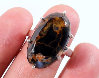 Op de afbeelding: Ovale ring met een gepolijste, donkere edelsteen met bruine en gouden adering. De steen is gezet in een zilveren band met kleine, decoratieve pinnen. De ring wordt door een vinger omhoog gehouden.