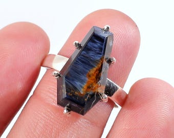 Op de afbeelding: Een zilveren ring met een rechthoekige, donkerblauwe edelsteen met bruine en gouden strepen. De edelsteen is gezet met kleine zilveren accenten. De ring wordt door een vinger omhoog gehouden.