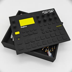 Könnte beinhalten: Schwarzes elektronisches Musikproduktionsgerät mit dem Text "SOULBRIX" und "Digibox". Das Gerät hat einen gelben Bildschirm und Knöpfe sowie eine Schublade mit kleinen goldfarbenen Objekten. Das Gerät befindet sich in einer schwarzen Box.