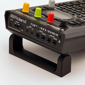 May include: A dark gray Roland SP-404MKII sampler featuring a black chassis, a grid of black rubber pads, and a central circular display. The device includes three colorful knobs in yellow, lime green, and red, plus a silver knob, mounted on a stand.