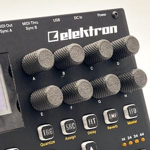 Puede incluir: Un muestreador de audio negro Elektron que presenta una cuadrícula de ocho perillas giratorias texturizadas y moleteadas etiquetadas de la A a la H. Debajo de las perillas hay botones cuadrados etiquetados TRG, SRC, FLT, AMP y LFO con texto amarillo. El panel superior incluye puertos MIDI, USB y DC In.