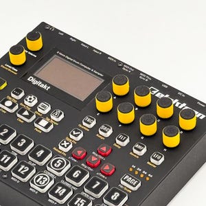 May include: A black Elektron Digitakt 8-voice digital drum computer and sampler featuring a central screen, numerous white backlit buttons with black numbering, and several yellow-topped rotary knobs arranged on the right side and top left.