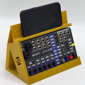 Op de afbeelding: Een mosterdgele 3D-geprinte standaard met een zwarte Zoom LiveTrak L6 digitale mixer en een donkere smartphone. De mixer is voorzien van diverse ingangen, draaiknoppen en knoppen met witte labels. De standaard heeft een driehoekig profiel.
