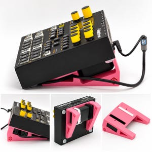 Pode incluir: Um sampler de música Elektron Digitakt preto montado num suporte de plástico cor-de-rosa brilhante inclinado. O dispositivo possui botões amarelos, teclas brancas e um cabo de alimentação preto. O suporte tem um design geométrico vazado.