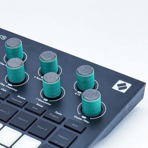 May include: A black music production controller featuring five textured teal knobs with grey tops. The device includes a grid of square buttons labeled with functions like Drum, Mutate, Duplicate, Save, Projects, Packs, Shift, and Patterns, plus a Master Filter label.