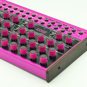 May include: A bright pink analog synthesizer labeled EDGE with a black control panel featuring numerous textured pink knobs, multiple toggle switches, and a grid of patch points. The device has dark side panels and a metallic pink chassis.
