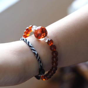 May include: Two bracelets are shown. One is a beaded bracelet with amber-coloured beads and a large, clear, amber-coloured bead. The other bracelet is black and white braided. The text "CRYSTAL MATRIX" is at the top.