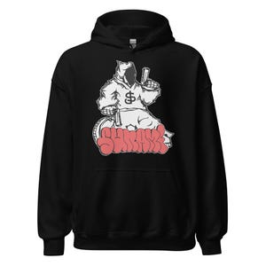 May include: Black hooded sweatshirt featuring a white graphic of a hooded figure holding a gun and a dollar sign. The word "SHRAKK" is written in red graffiti-style lettering across the bottom of the graphic.