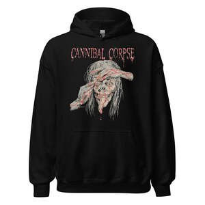 Puede incluir: Sudadera con capucha negra con el nombre de la banda "CANNIBAL CORPSE" en fuente roja desgastada. Debajo, un gráfico de una cara de zombi con carne expuesta y pelo largo y blanco. El gráfico es detallado y grotesco.