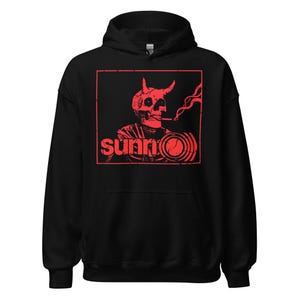 May include: Black hooded sweatshirt featuring a red graphic of a horned skull smoking a cigarette, with the word "sunn" in a red, stylized font. The design is enclosed in a red rectangular frame. The sweatshirt has a drawstring hood and a front pocket.