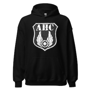 May include: Black hooded sweatshirt featuring a distressed white graphic. The graphic is a shield shape with the letters "AHC" at the top, wings, and a starburst design. The sweatshirt has a drawstring hood and a front pocket.