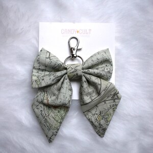 May include: A bow-shaped keyring with a map design in shades of grey and green. The keyring has a silver-coloured clasp and ring. The tag reads "CANDY + CULT".