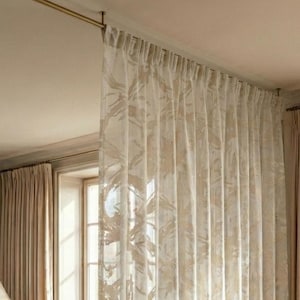 May include: Cream-coloured, patterned sheer curtains hang from a gold-toned rod, partitioning a living space. A brown velvet sofa, marble coffee table, and dining table with chairs are also visible.