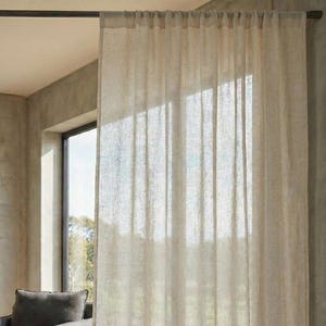 May include: A sheer, light beige curtain hangs in front of a window, casting soft shadows. The curtain is made of a textured fabric, and the sunlight filters through, illuminating the room. A gray armchair and wooden coffee table are visible.