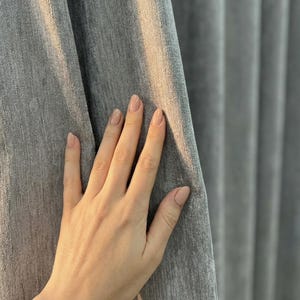 Könnte beinhalten: Eine Hand mit manikürten Nägeln berührt einen grauen Vorhang. Der Vorhang hat ein weiches, strukturiertes Aussehen mit vertikalen Falten. Die Beleuchtung erzeugt Schatten und Glanzlichter auf dem Stoff und betont seine Textur und Farbe.