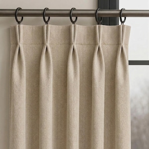 May include: Beige herringbone patterned curtain panel hanging from a dark metal rod with black rings. The curtain is pleated at the top, creating vertical folds. The fabric has a subtle texture and a neutral color, suitable for various interior styles.