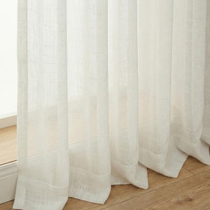 May include: Off-white linen curtains hanging in front of a window. The curtains have a textured weave and are partially translucent, allowing light to filter through. The bottom hem of the curtains is visible, and the floor is made of wood.
