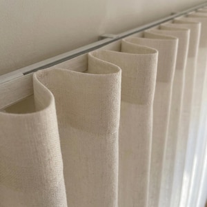 May include: Off-white linen curtains featuring a structured S-fold heading attached to a white metal track. The fabric has a visible woven texture and hangs in uniform, vertical waves, creating a clean and consistent pleated appearance.