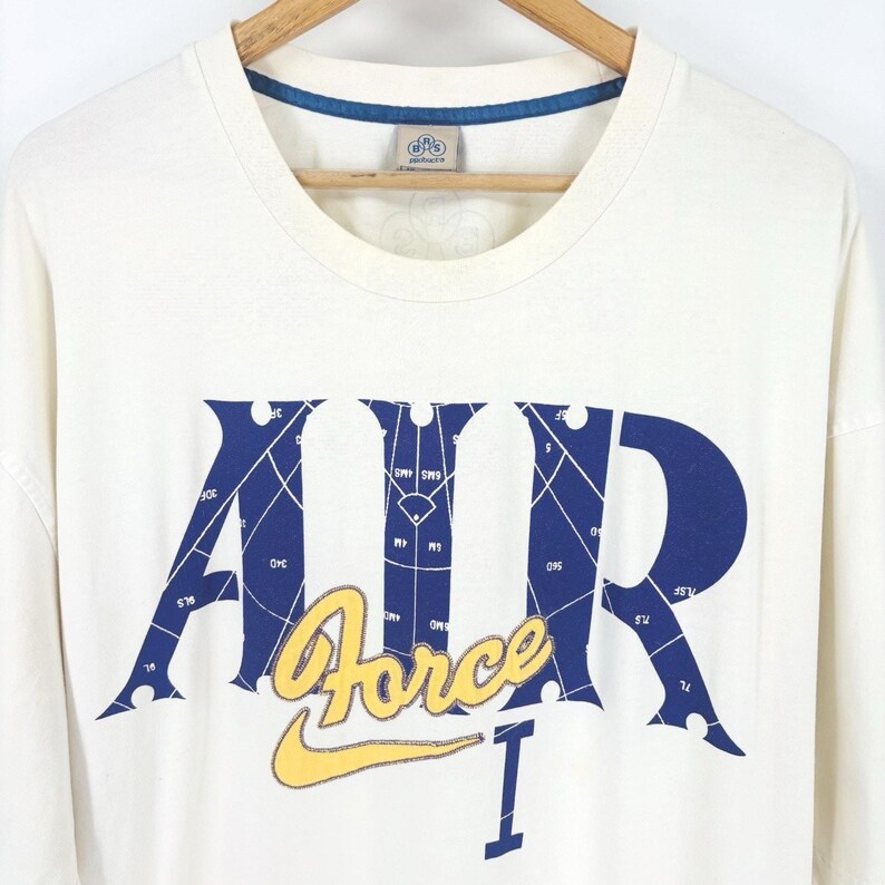 May include: Cream-colored long-sleeve t-shirt with a blue and yellow graphic. The graphic features the word "AIR" in blue with a baseball field design, the word "Force" in yellow, and the number "1" in blue.