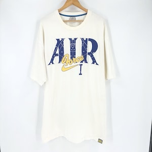 May include: Cream-colored t-shirt with the words "AIR Force I" in navy blue and yellow. The Nike logo is visible below the word "Force". The shirt is hanging on a wooden hanger.