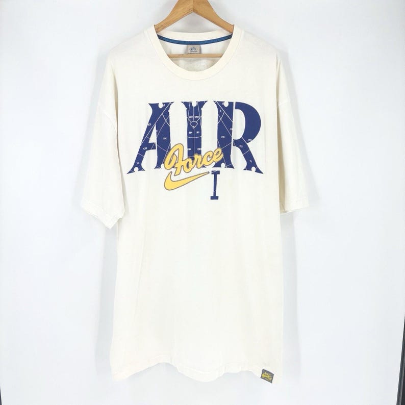 May include: Cream-colored t-shirt with the words "AIR Force I" in navy blue and yellow. The Nike logo is visible below the word "Force". The shirt is hanging on a wooden hanger.