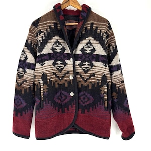 Peut inclure: Veste boutonnée à motifs géométriques dans les tons de marron, noir, violet et bordeaux. La veste a un col cranté, deux poches avant et trois boutons argentés. Elle est suspendue à un cintre en bois.