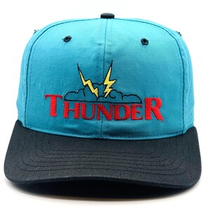 Pode incluir: Um boné de beisebol azul-petróleo com aba preta e um design bordado na frente. O bordado inclui uma nuvem cinza com dois raios amarelos acima e a palavra THUNDER costurada em letras maiúsculas vermelhas em destaque na parte inferior.