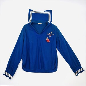 Peut inclure: Une chemise de marin bleue vintage avec un col et des poignets bordés de blanc. La chemise à manches longues présente des écussons brodés sur la poitrine, notamment des étoiles et un emblème circulaire. Le tissu est léger.