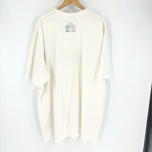 May include: Cream-colored t-shirt hanging on a wooden hanger. The back of the shirt features a small black graphic of three connected circles with the word "proctes" below. The shirt has short sleeves and a crew neck.