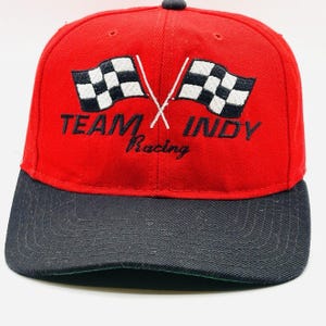May include: A red baseball cap with a black brim featuring an embroidered design of two crossed checkered racing flags. The text TEAM INDY Racing is stitched in black thread across the front panel, positioned below the racing flags.